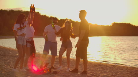Three-girls-and-two-boys-spend-summer-evening-at-sunset-on-the-beach-in-shorts-and-t-shirts-around-bonfire-with-beer.-They-are-clink-beer-and-dancing-on-the-open-air-party.